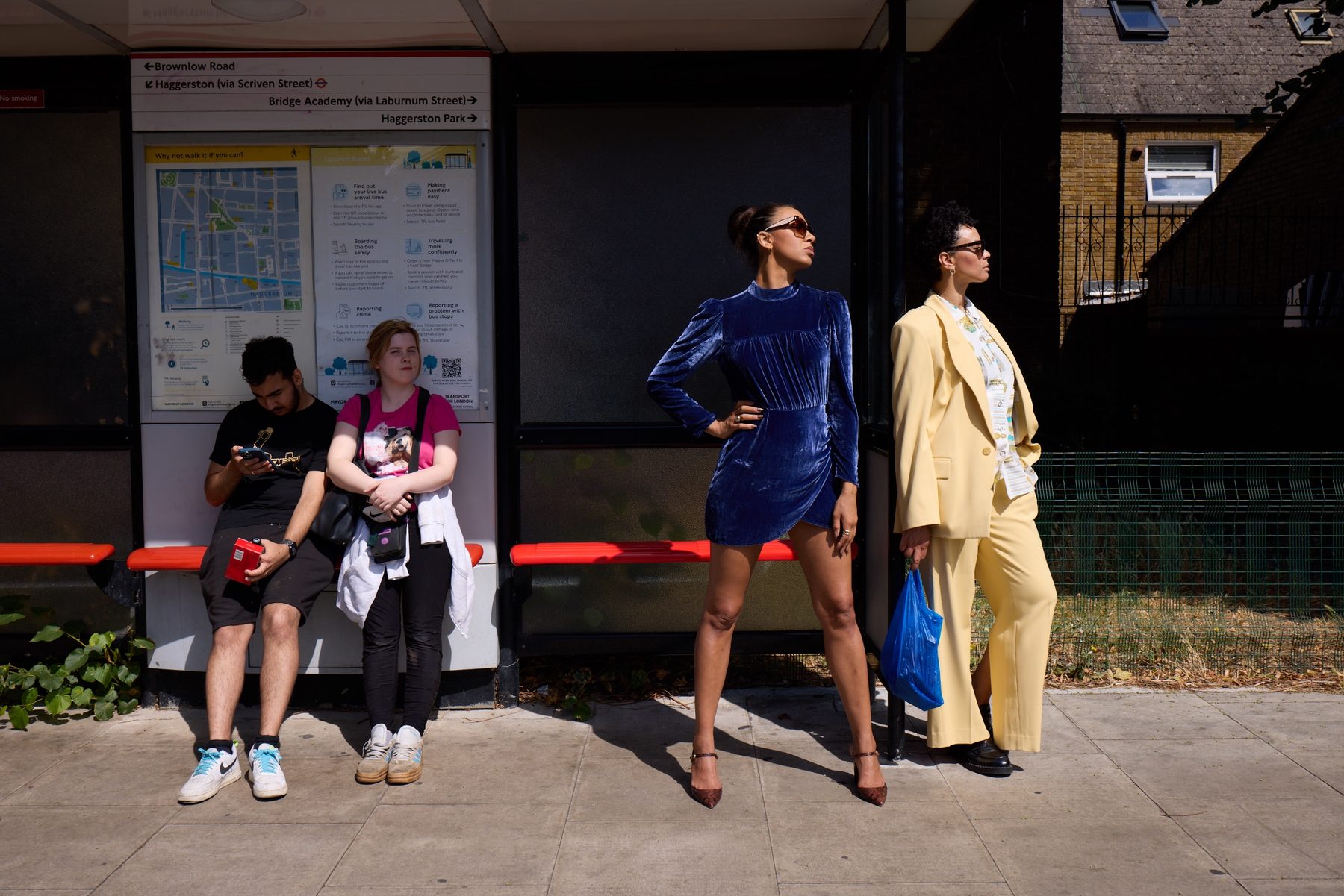 Mel in blue velvet and Holly in a pale yellow suit posing at a London bus stop